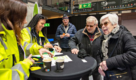 Två kvinnor i gul varseljacka ler och lämnar biljetter till en äldre dam och två herrar.