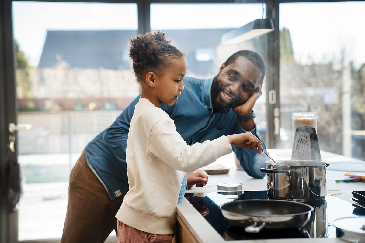 En vit köksö med spishäll, där en kastrull med vatten kokar. En man i blå skjorta står lutad mot köksön och tittar på när en flicka rör om i grytan.