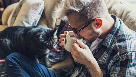 Vuxen person sitter i soffan och dricker te med katten i knä.
