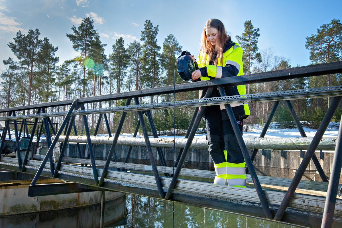 En kvinna i varselkläder står på en bro över en bassäng med ett verktyg för att mäta vattennivån.