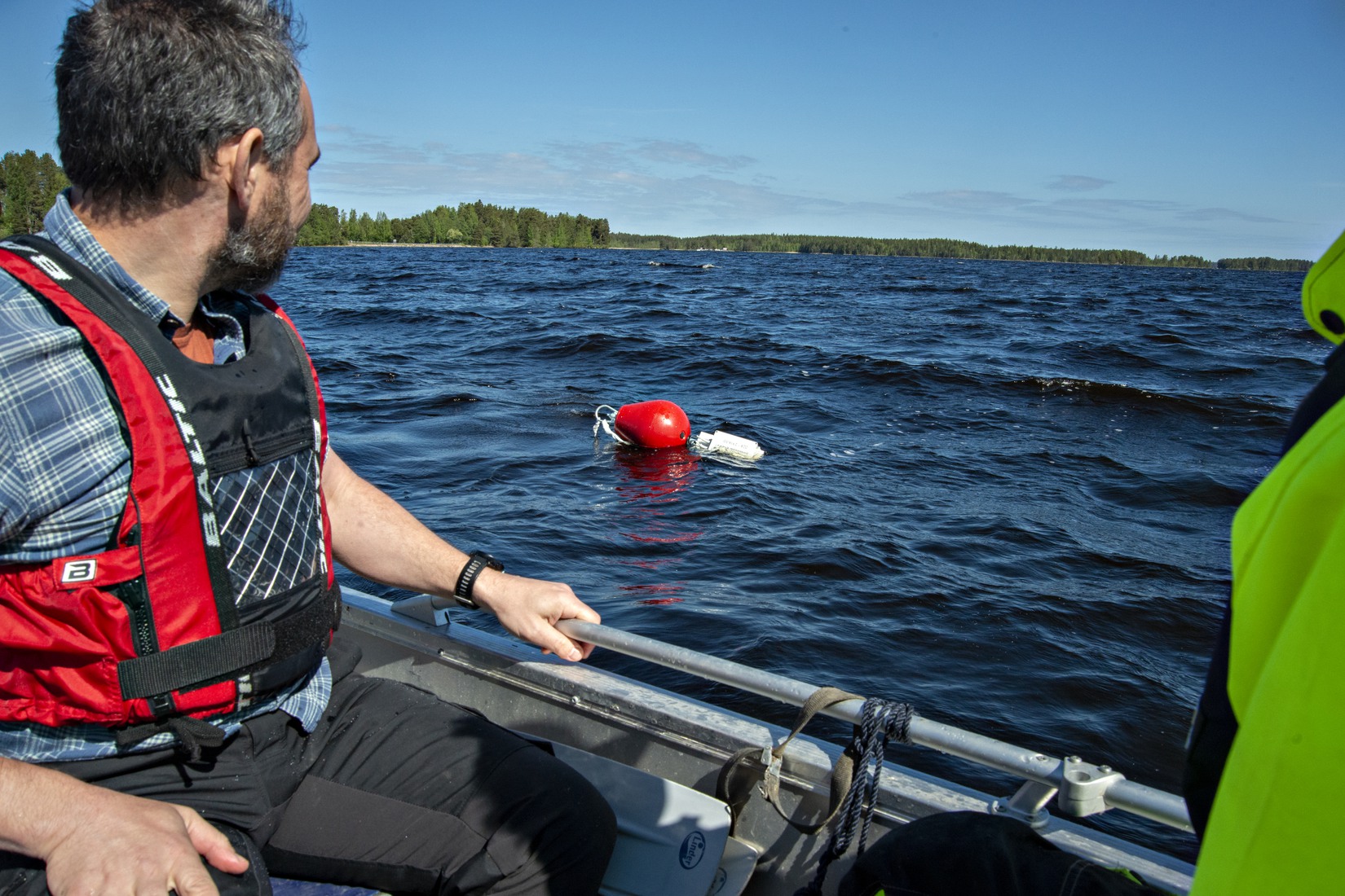 En röd boj syns i sjön. Två personer i båt i förgrunden.